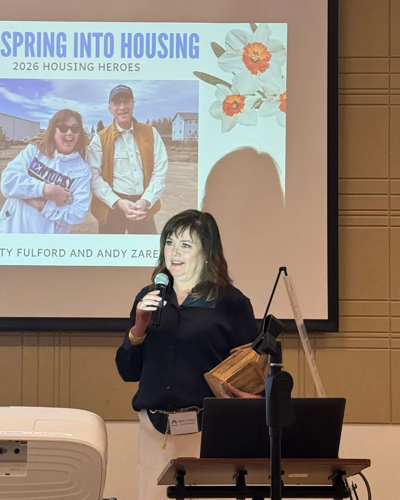 Marty Fulford speaking at the Corvallis Housing First Spring Into Housing event, holding a wooden award, with the 2026 Housing Heroes slide behind her